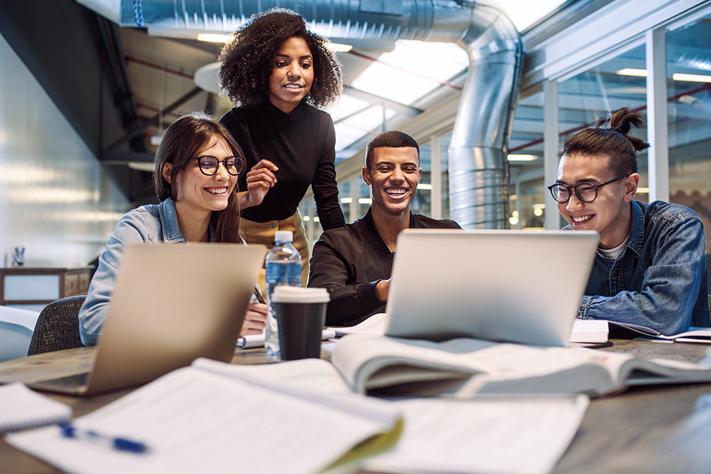 A diverse group of young adults collaborates around laptops, notebooks, and coffee, exemplifying Managed Learning Services in action.