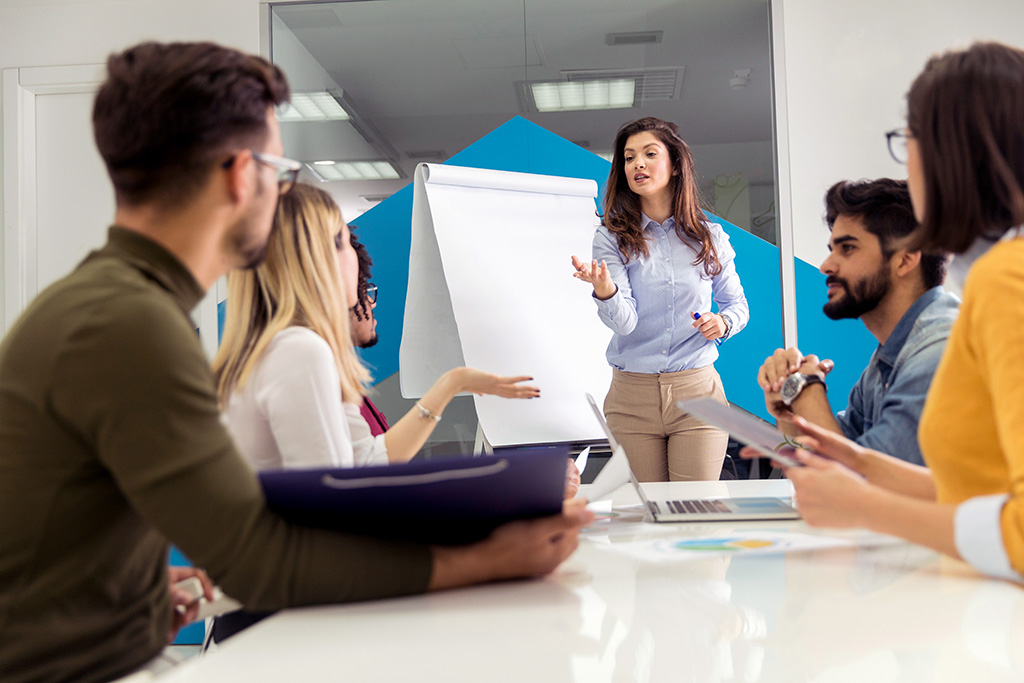 A woman leads a discussion with a group around a table, using a flipchart, demonstrating Managed Learning Services in a meeting setting.
